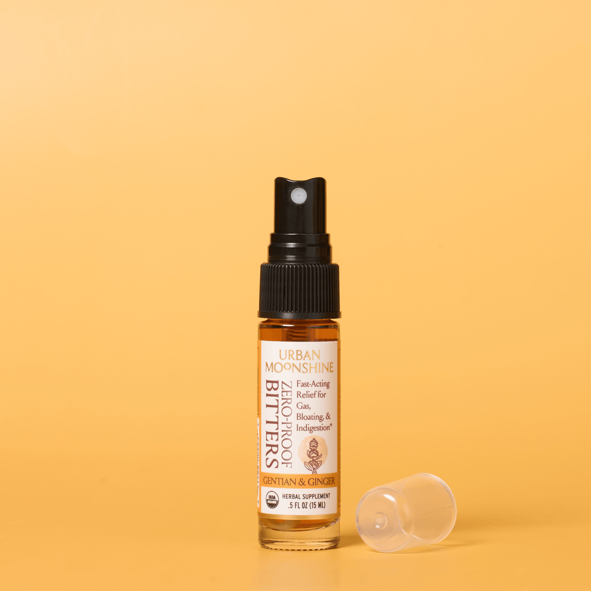 A small amber glass spray bottle labeled Urban Moonshine Bitters with a black pump, standing on a warm yellow background with its clear cap placed beside it.