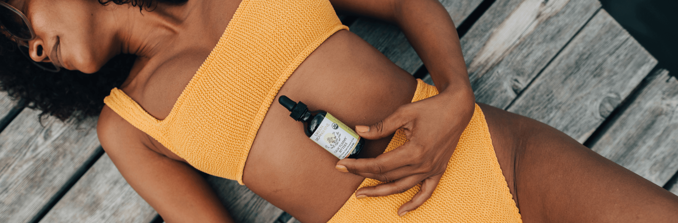 A woman in a yellow ribbed crop top and matching bottoms lies on weathered wooden planks, holding a small dropper bottle against her stomach.
