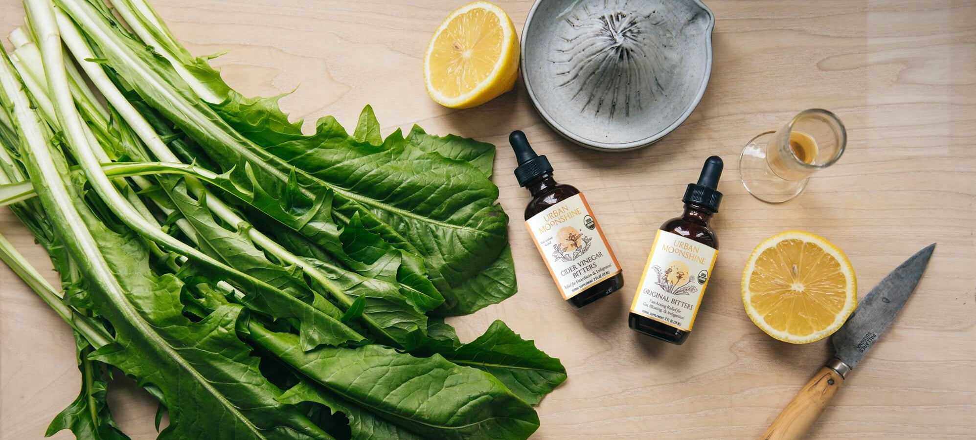 Bunch of green leafy vegetables on a wooden surface with bitters bottles, lemons, and a knife.