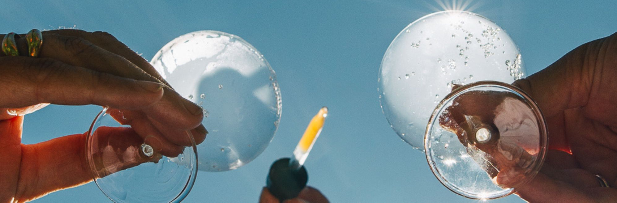 Two hands holding glasses with bubbles and a tincture dropper against a clear blue sky.