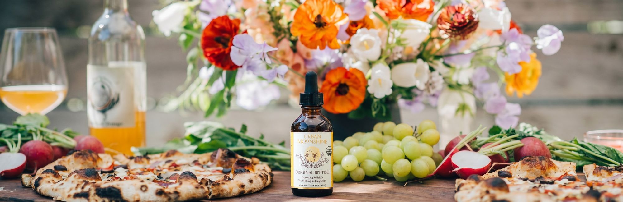 Bottle of bitters with a bouquet of flowers and pizza on a wooden table
