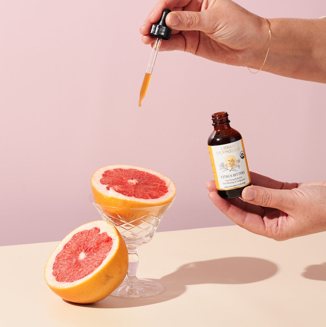 Hand holding a bottle of citrus bitters with a dropper over a halved grapefruit on a pink background.