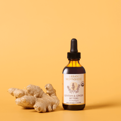 A bottle of Urban Moonshine Gentian & Ginger Bitters next to fresh ginger roots on a warm yellow background.