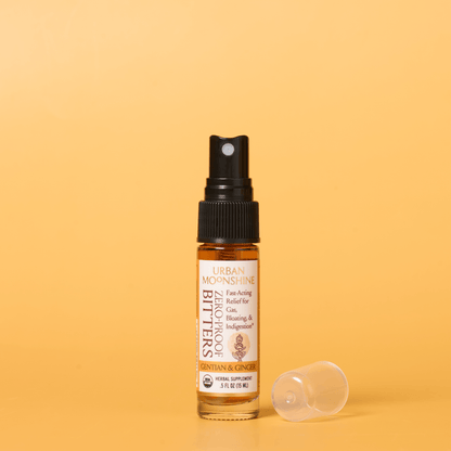 A small amber glass spray bottle labeled Urban Moonshine  Bitters with a black pump, standing on a warm yellow background with its clear cap placed beside it.