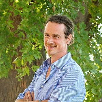 Guido Mase wearing a blue striped shirt standing outdoors with greenery in the background