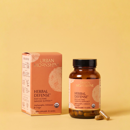 Urban Moonshine Herbal Defense dietary supplement shown with its orange box and amber bottle on a solid yellow background, with two capsules lying beside the bottle.