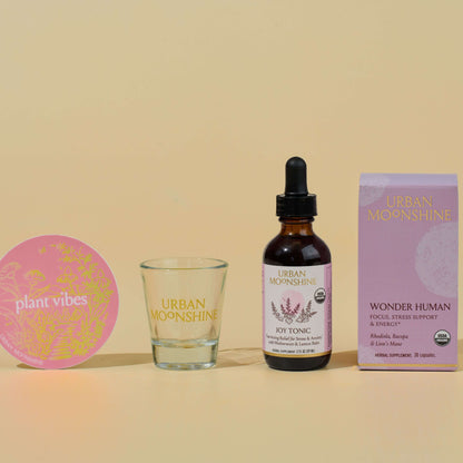 Bottle of Urban Moonshine Joy Tonic with a glass, pink coaster, and box on a beige background