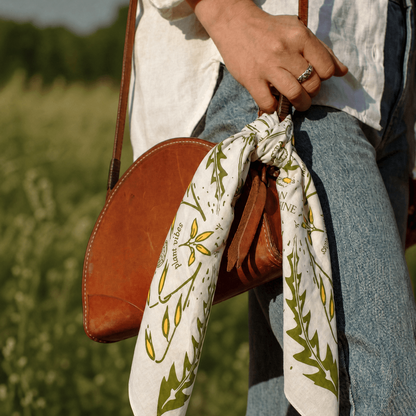 Person holding a brown leather bag with a floral bandana draped over it, standing in a field.